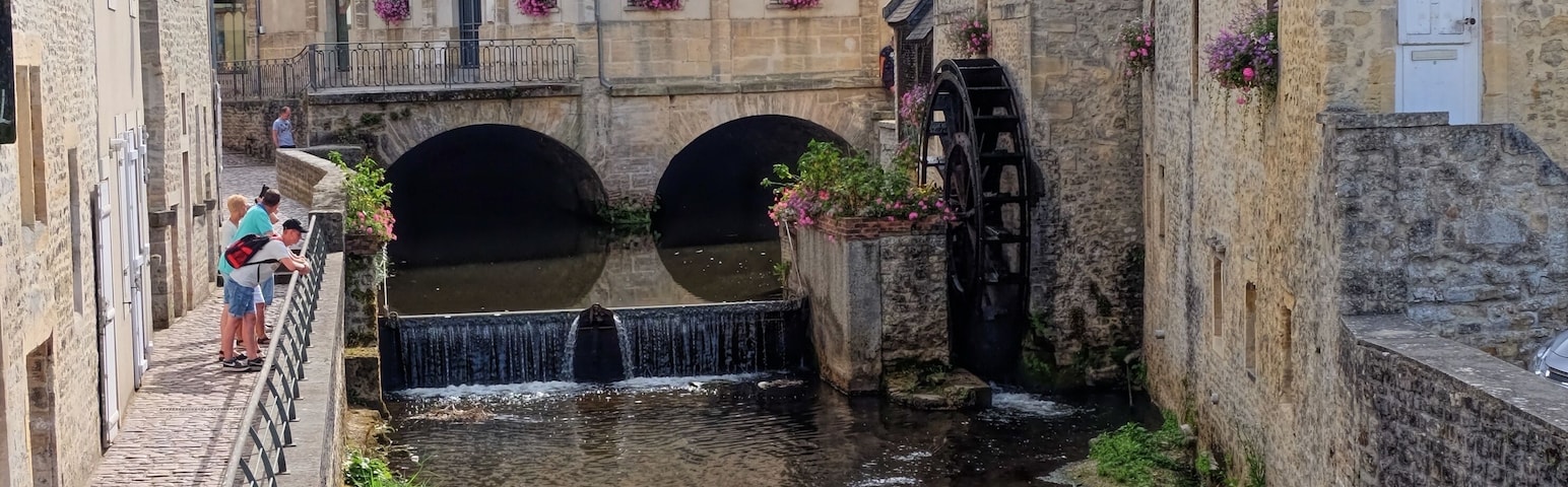 Un canal estrecho con un molino de agua y un puente, rodeado de edificios históricos de piedra y la aguja de una iglesia al fondo.