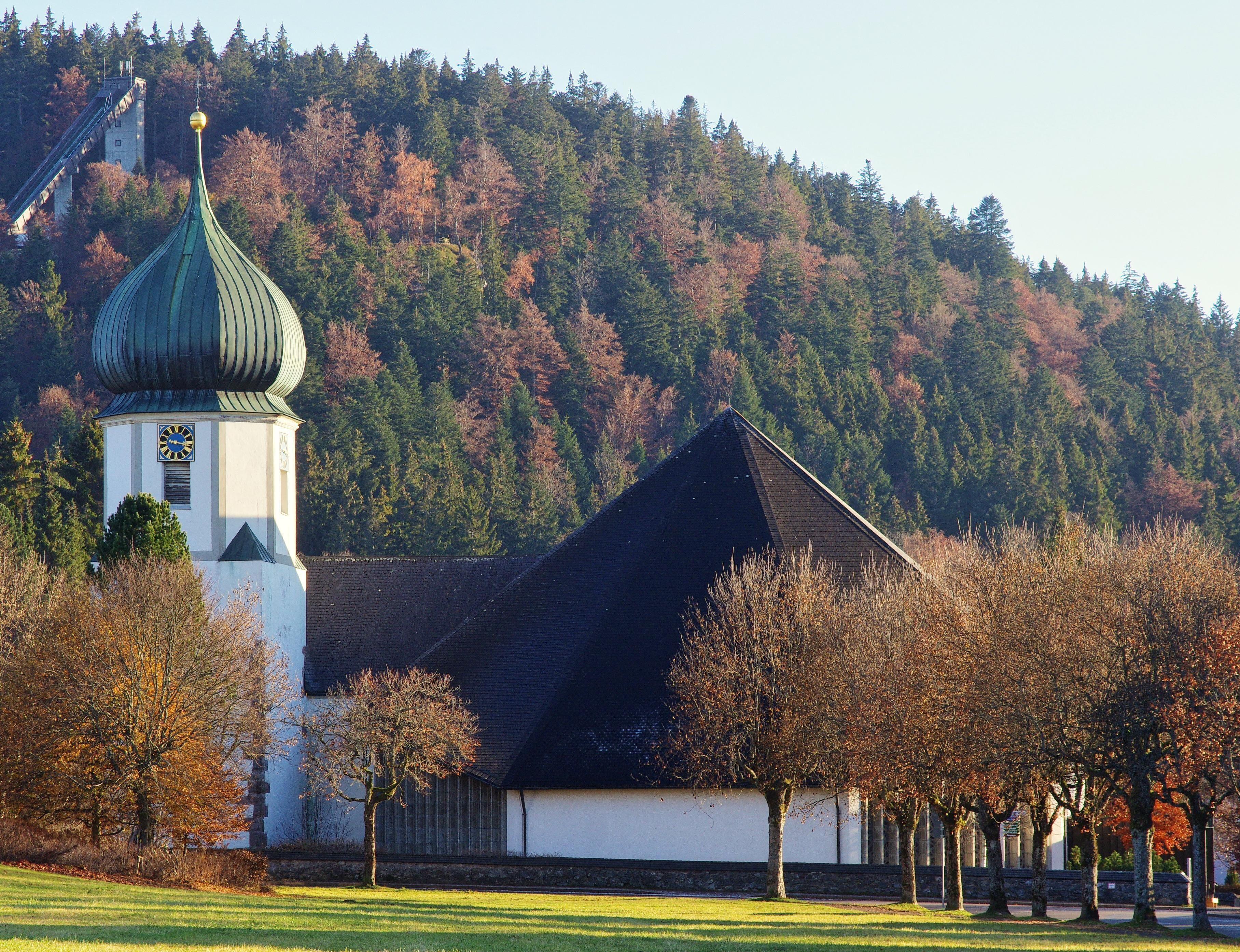 A church with a green dome and a clock tower, surrounded by trees and a hill in the background.