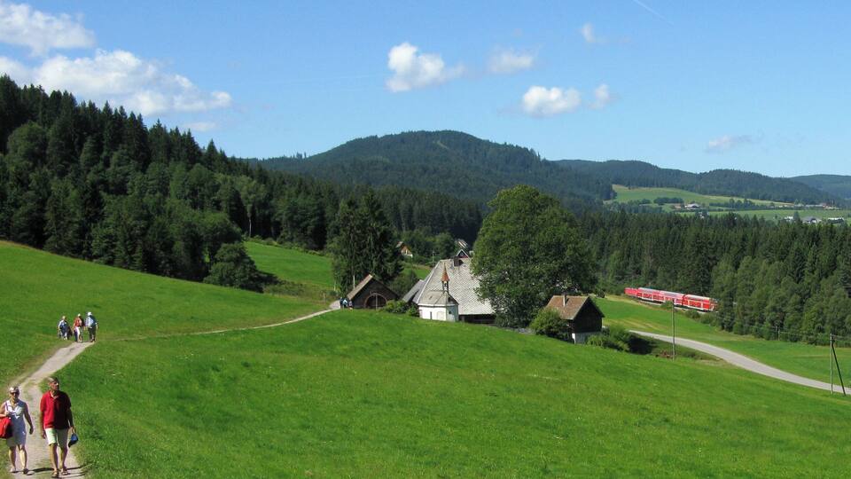 A green valley with a small church, a red train, and hikers on a path.