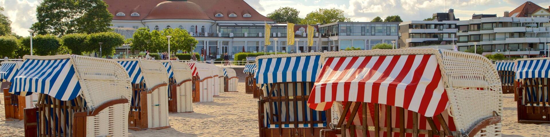 Beach huts with striped awnings, sandy beach, and a building with a red roof in the background.