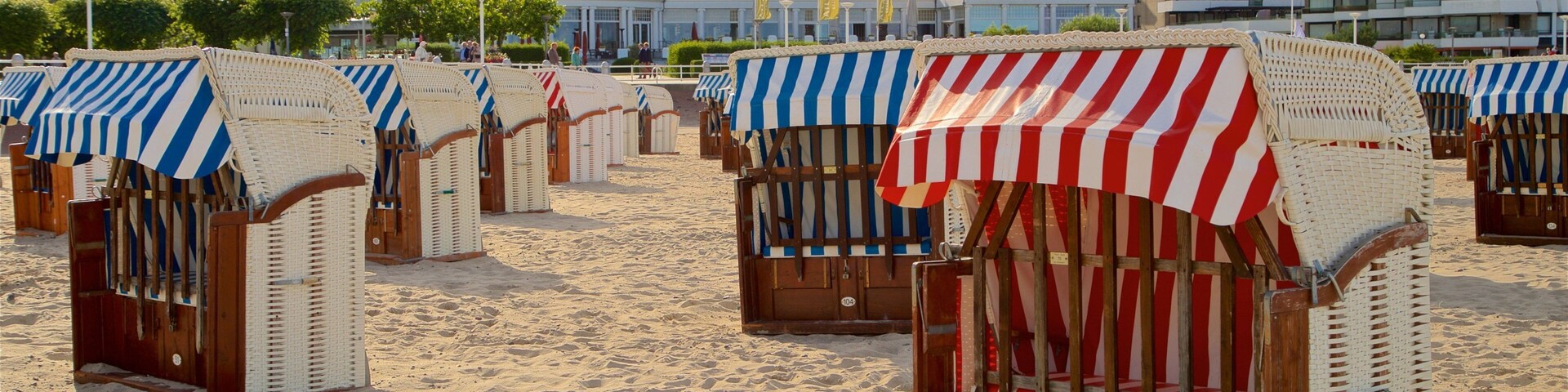 Cabañas de playa con toldos a rayas, playa de arena y un edificio con techo rojo al fondo.