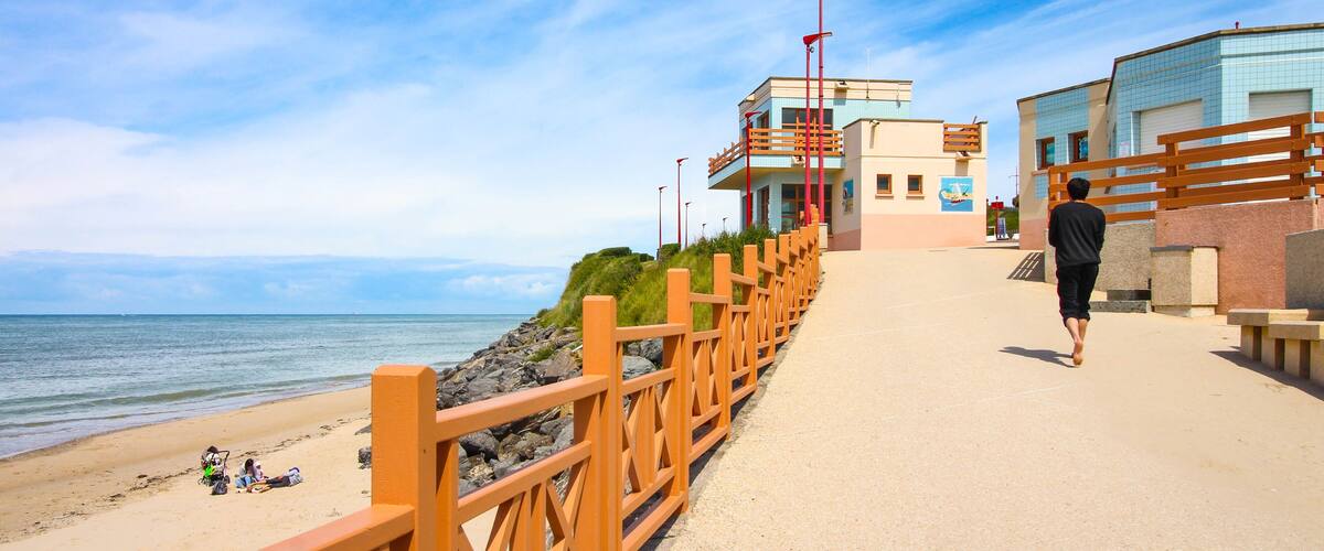 A coastal walkway with a wooden railing, beach, and a building with a blue and beige facade.