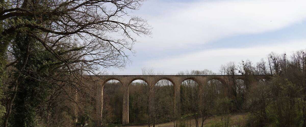 A stone viaduct over a river, surrounded by trees and a dirt path.