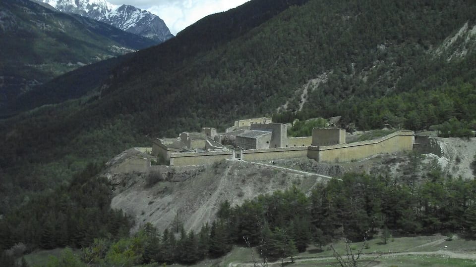 A mountainous landscape with a fortified structure surrounded by dense forests and snow-capped peaks in the background.