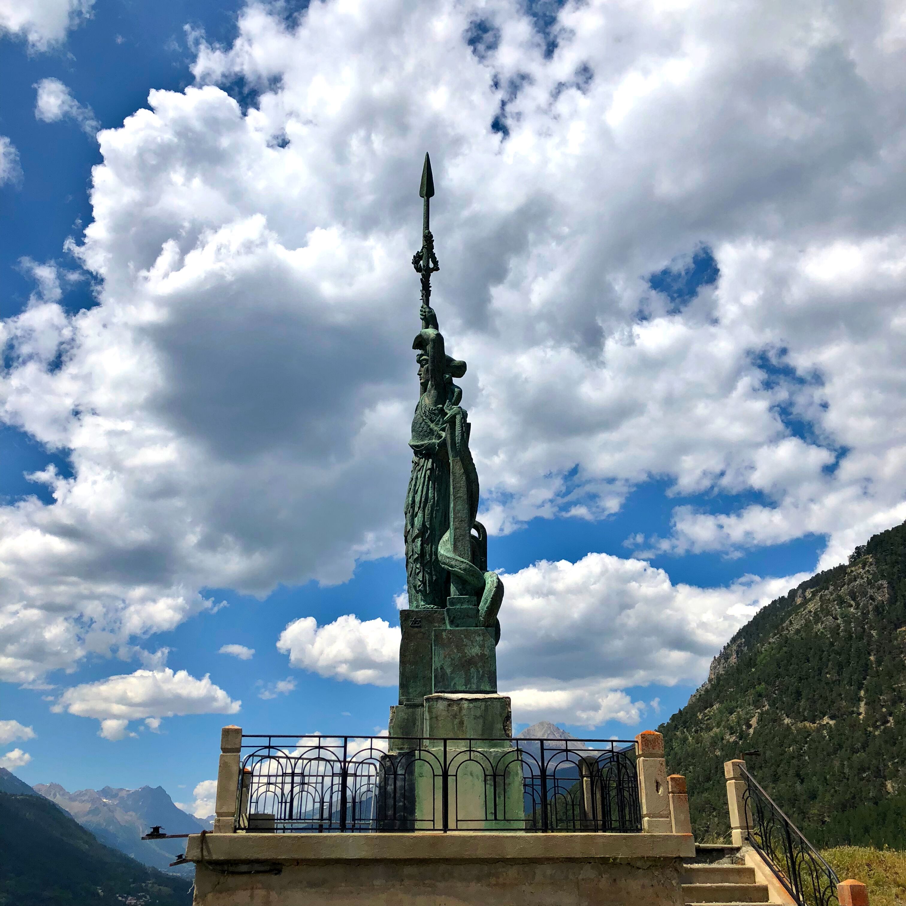 A statue of a figure holding a spear, set against a backdrop of mountains and a partly cloudy sky.