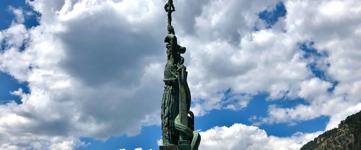 A statue of a figure holding a spear, set against a backdrop of mountains and a partly cloudy sky.