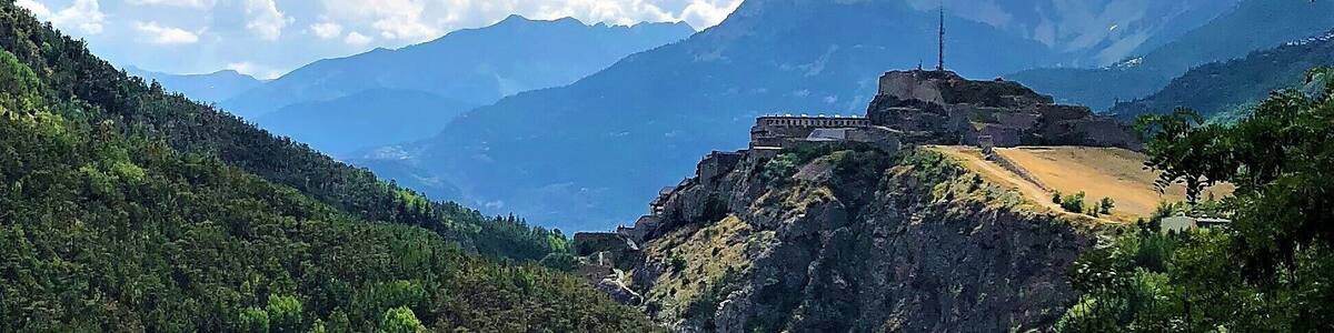 Un paysage montagneux avec un château imposant perché sur une colline, entouré d’une végétation luxuriante et surplombé d’un ciel bleu clair parsemé de quelques nuages.
