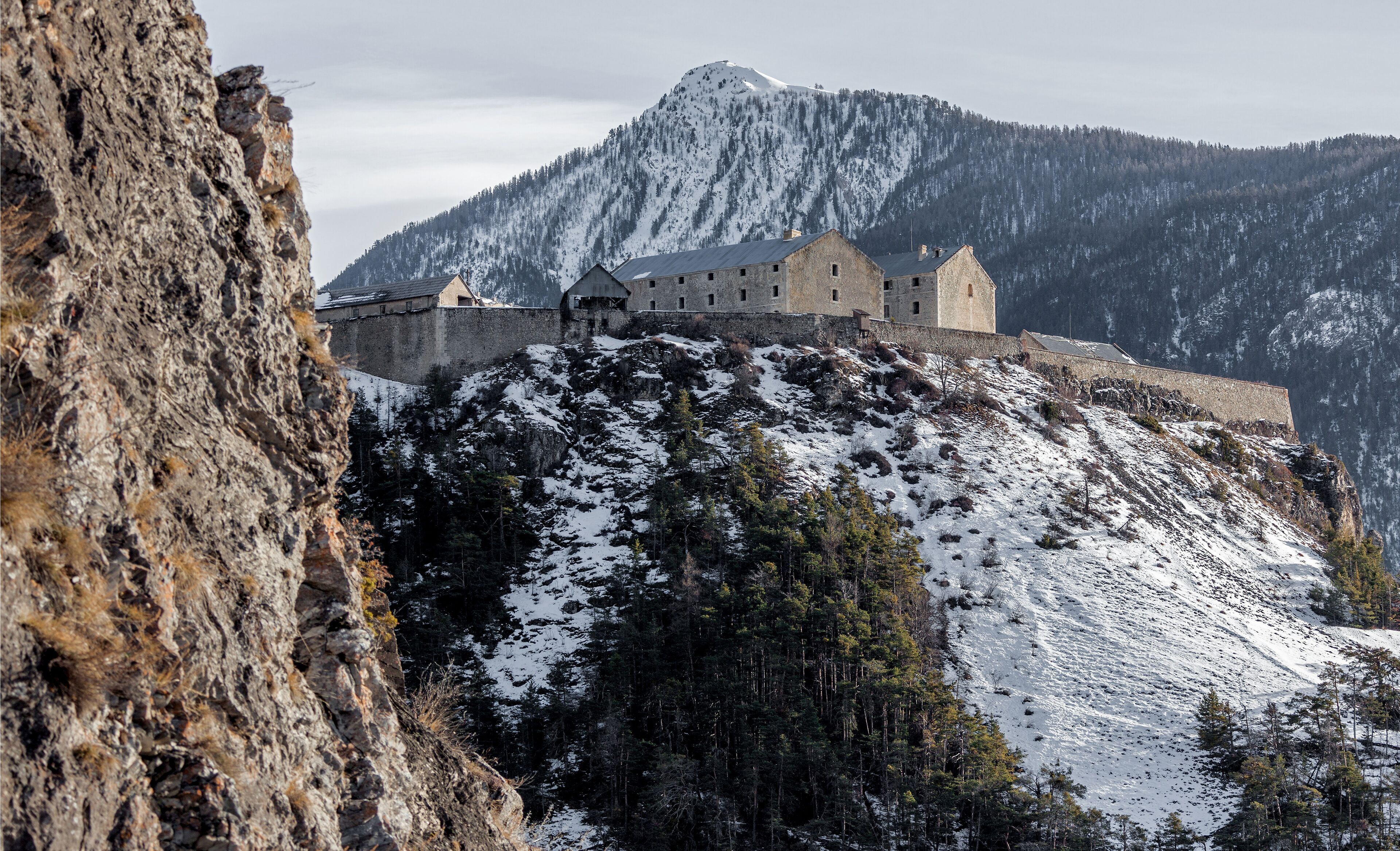A historic stone castle on a snow-covered mountain with a forested backdrop.