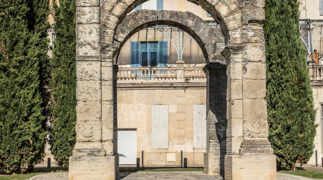 Un antiguo arco de piedra con un edificio moderno al fondo, rodeado de cipreses y un paisaje montañoso.