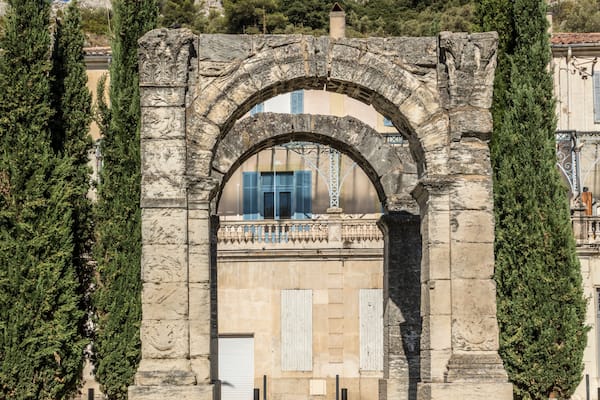 Un arc en pierre d’époque, avec un bâtiment moderne en arrière-plan, le tout entouré de cyprès et dominant un paysage montagneux.