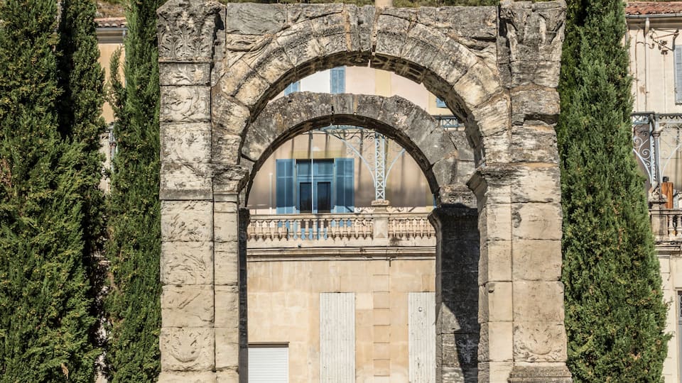 An ancient stone archway with a modern building in the background, surrounded by cypress trees and a mountainous landscape.