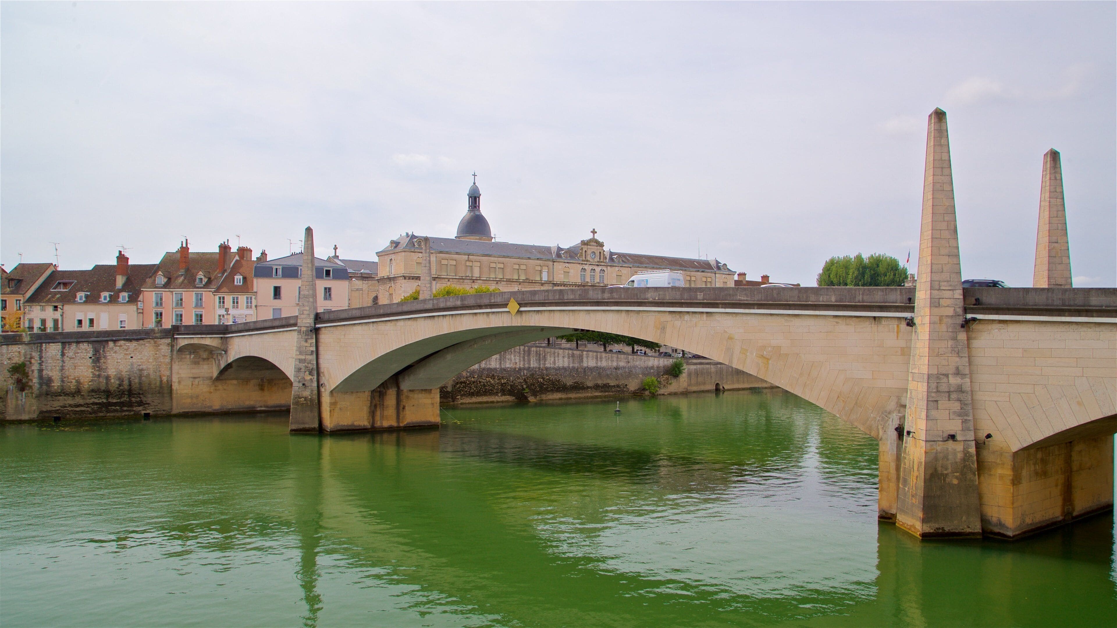 A stone bridge over a green river with buildings and a church in the background.