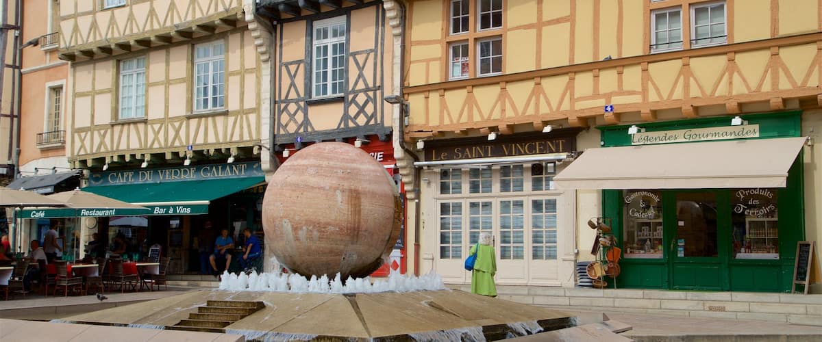 Una plaza tradicional de pueblo europeo con una gran fuente de piedra, edificios con entramado de madera y diversas tiendas.