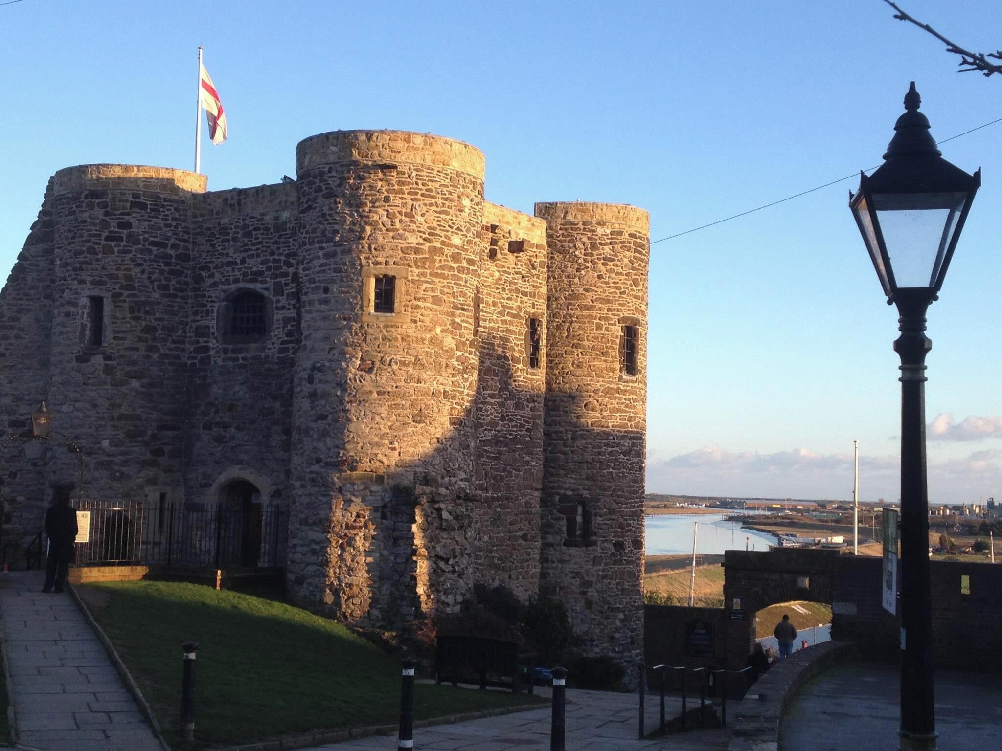 A historic stone castle with a flag, a street lamp, and a view of a body of water in the background.
