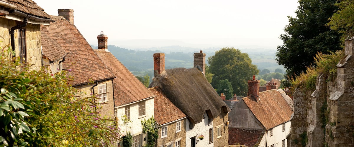 A cobblestone street lined with historic stone buildings, a thatched-roof house, and a stone wall.