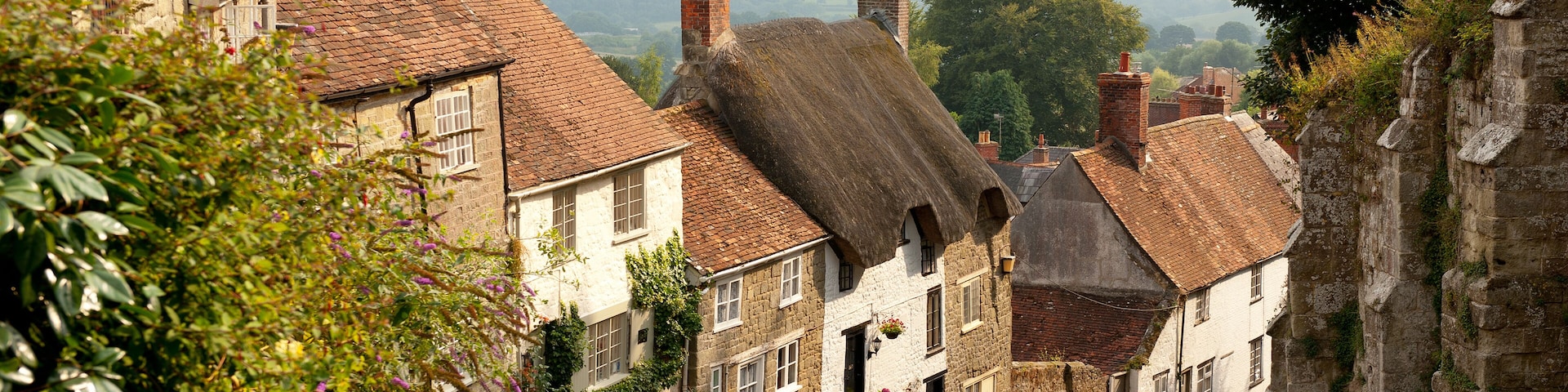 A cobblestone street lined with historic stone buildings, a thatched-roof house, and a stone wall.