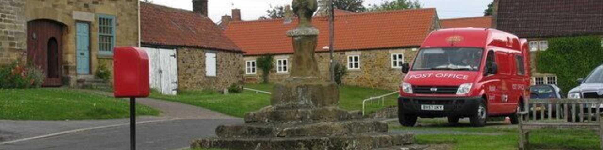 A stone fountain, a red post office van, and stone buildings in a village.