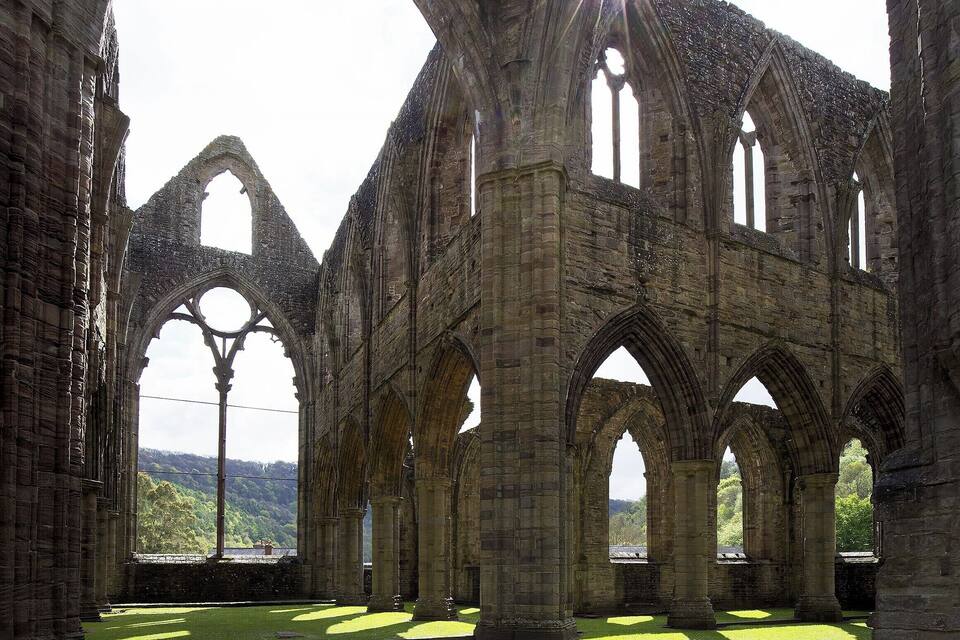 The ruins of a medieval stone structure with large arches and columns, set against a backdrop of lush greenery and a distant view of a bridge.