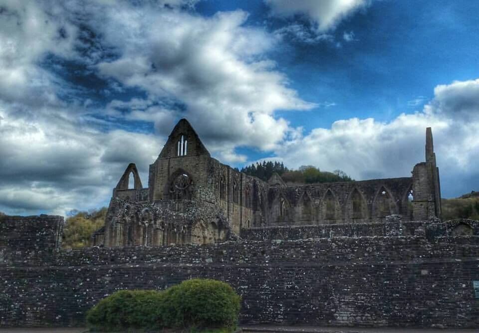 An ancient stone church with arched windows and a tall spire, surrounded by a stone wall and under a partly cloudy sky.
