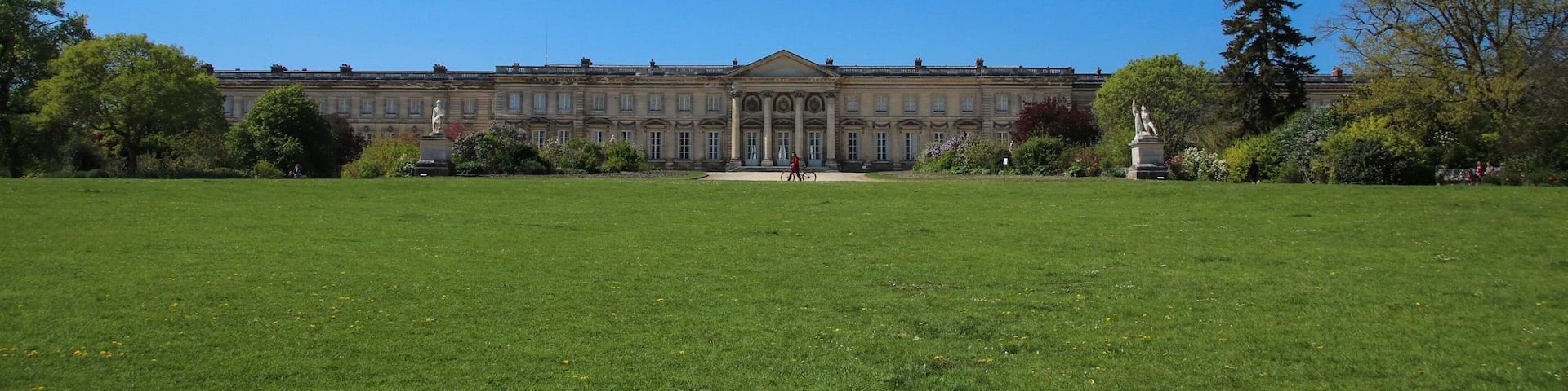 A large building with classical architecture, a well-maintained lawn, and statues in a sunny outdoor setting.
