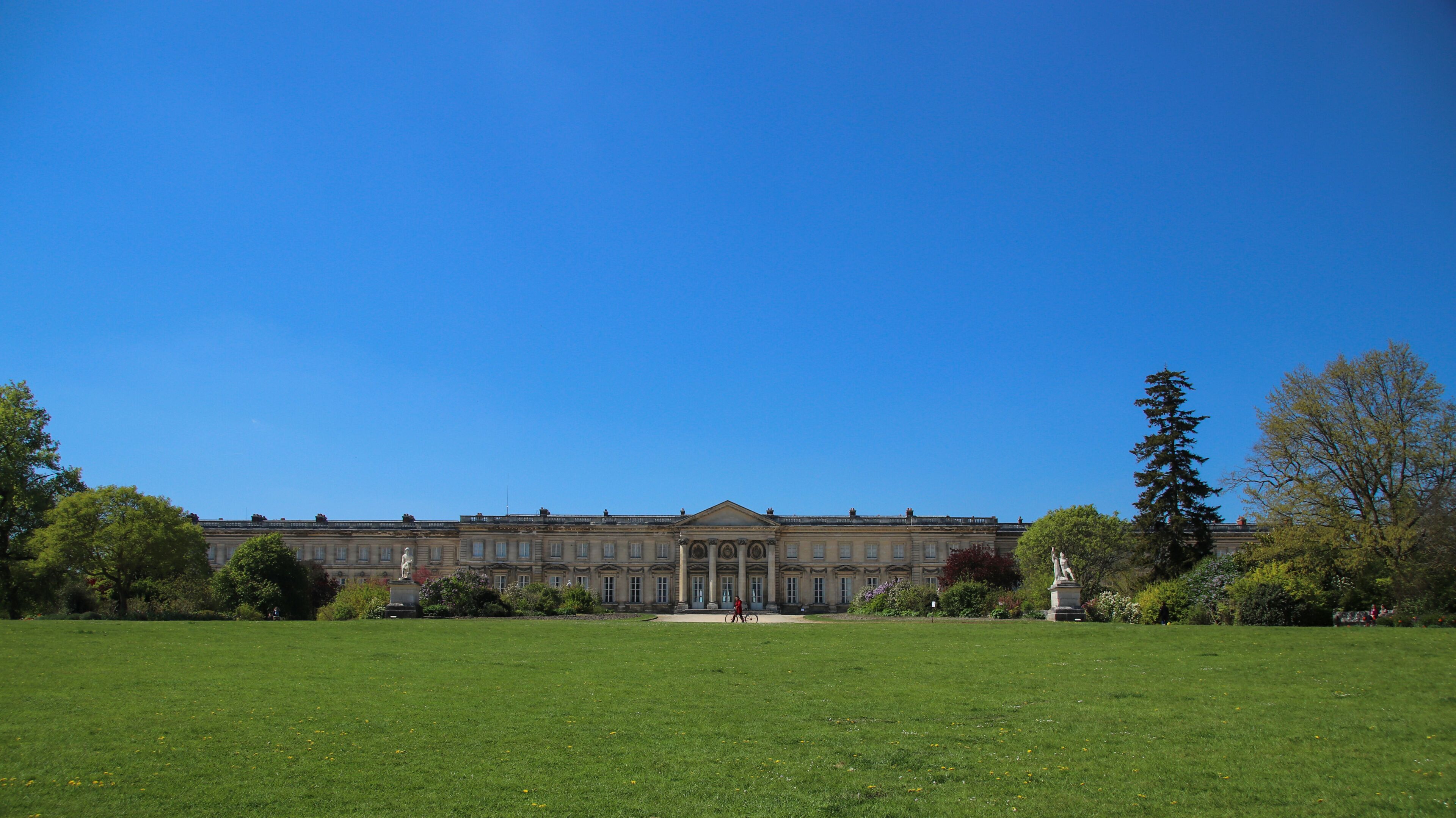 A large building with classical architecture, a well-maintained lawn, and statues in a sunny outdoor setting.