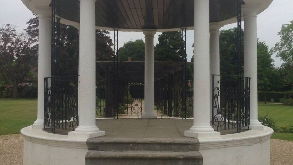 A white gazebo with a dome roof and columns, surrounded by gravel and greenery.