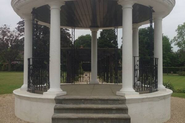 A white gazebo with a dome roof and columns, surrounded by gravel and greenery.