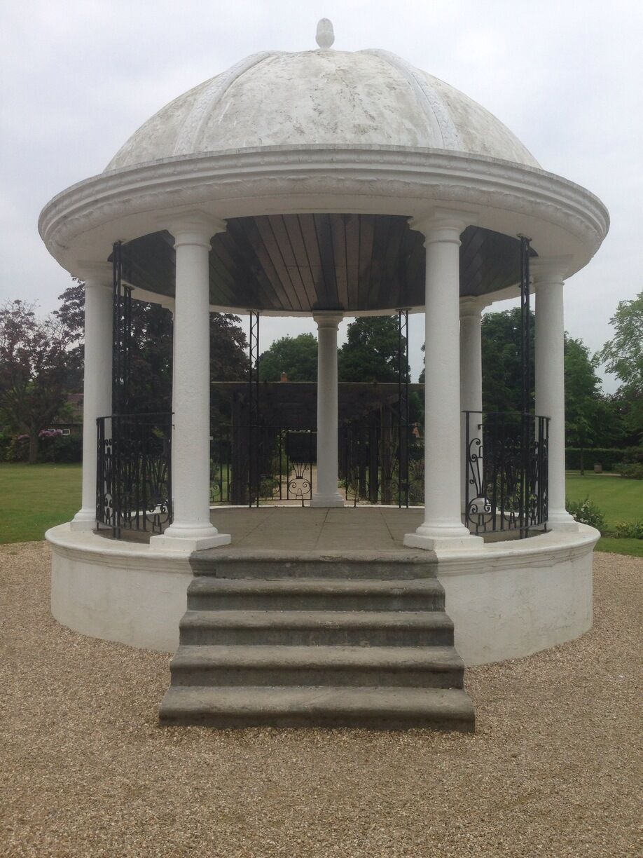 A white gazebo with a dome roof and columns, surrounded by gravel and greenery.