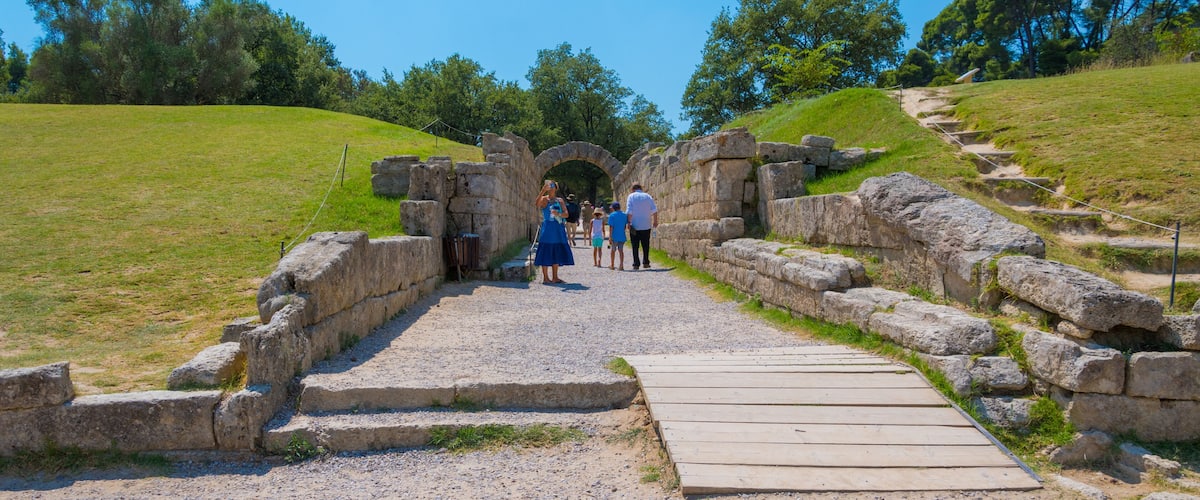 A historical stone archway leading to a grassy hill with a wooden pathway.