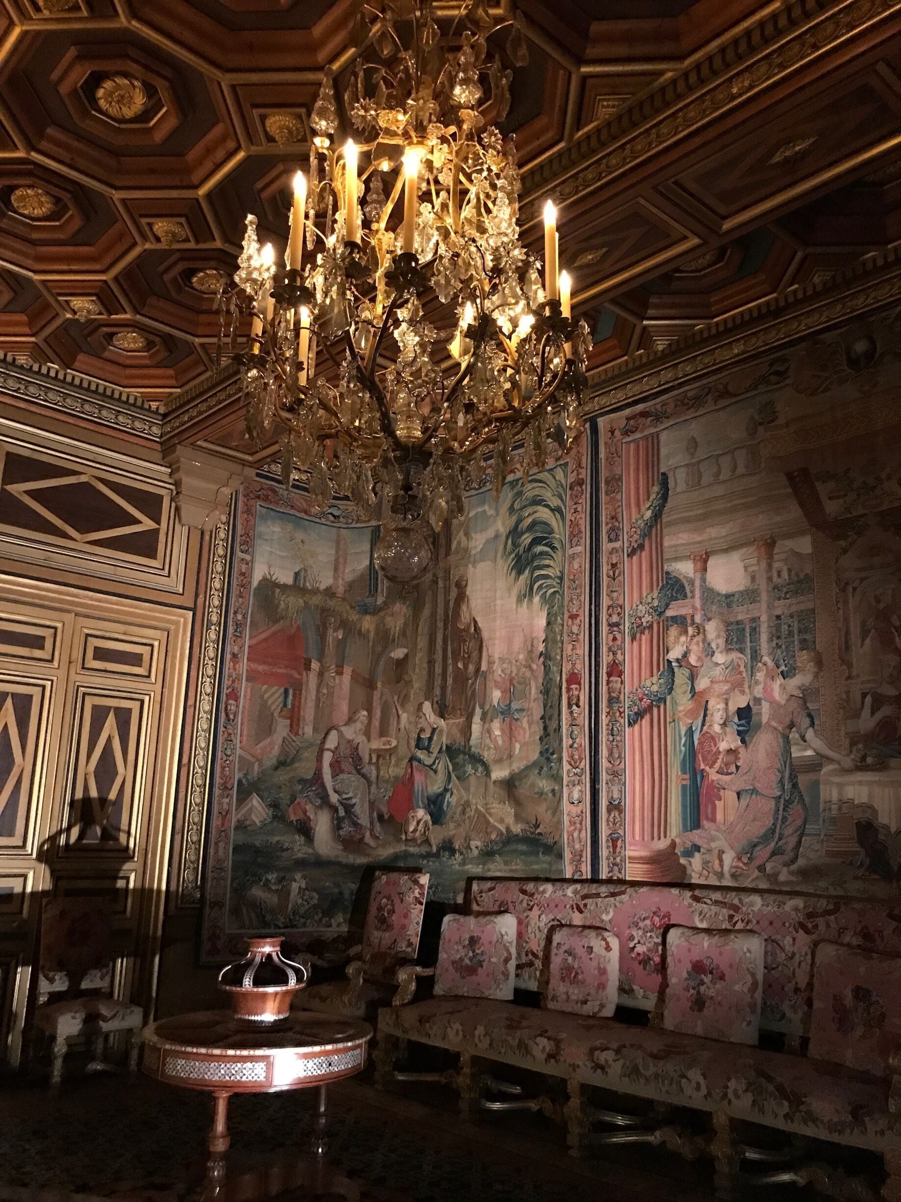 Ein reich verzierter Raum mit einem großen Kronleuchter, einem Wandgemälde und einem Sofa mit floralem Bezug.