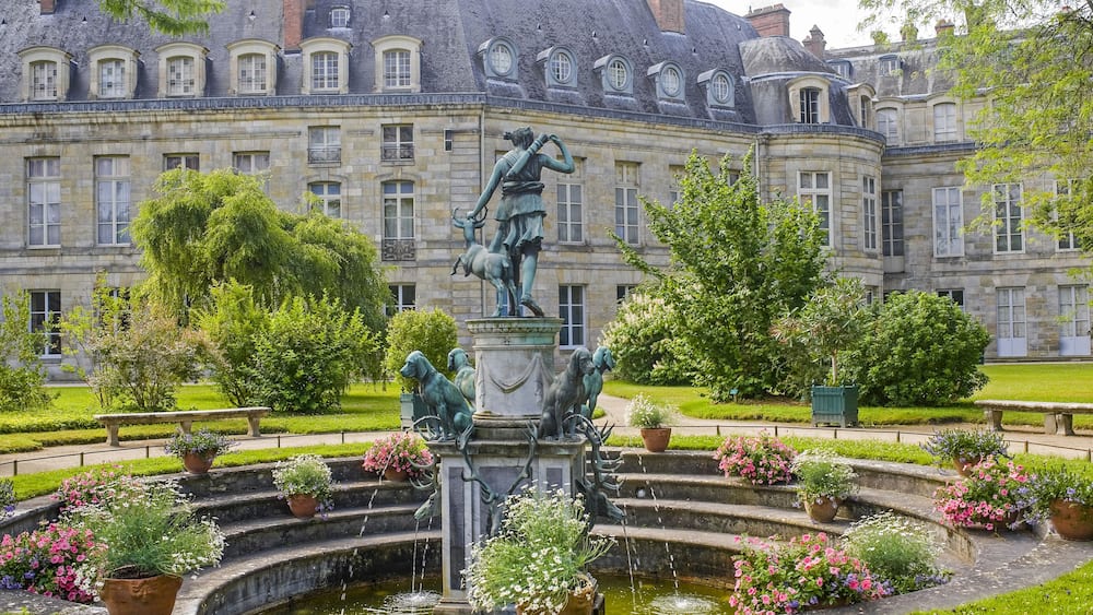 Un bâtiment historique doté d’une fontaine et de statues, situé dans un jardin bien entretenu.