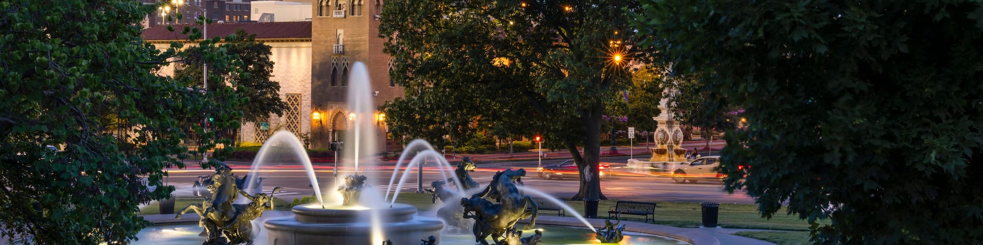 A cityscape with a prominent tower, a fountain with statues, and surrounding trees.