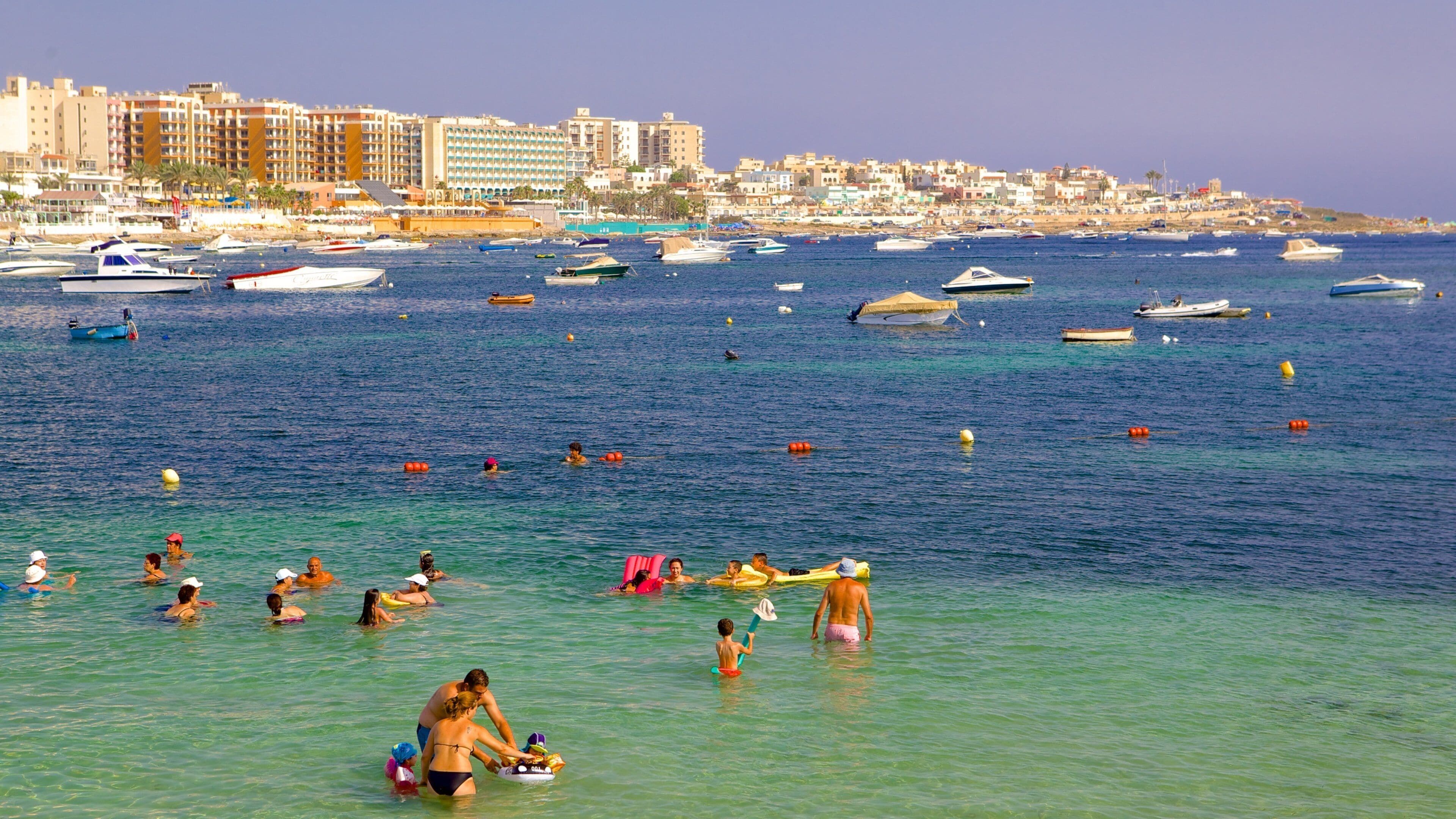 Una città costiera con un porto turistico pieno di imbarcazioni, persone che nuotano e si abbronzano in acqua, sullo sfondo edifici residenziali.