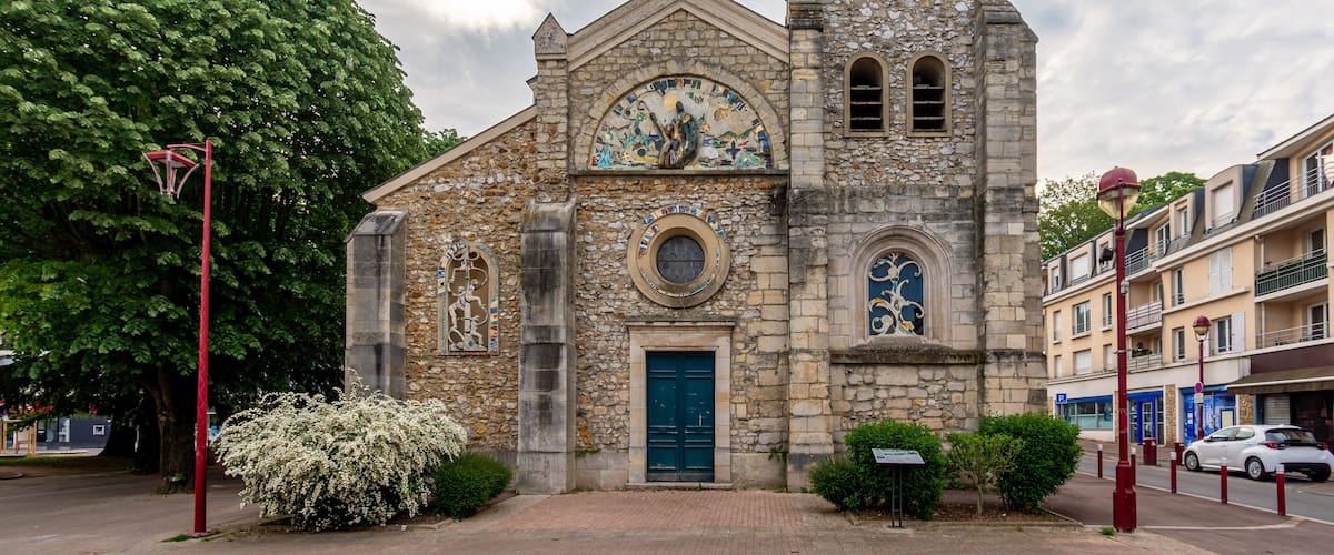 A historic stone church with a clock tower, surrounded by greenery and modern buildings.