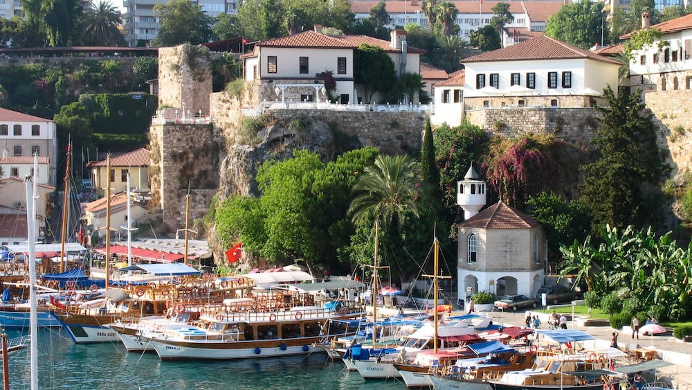 Una marina con imbarcazioni ormeggiate, un piccolo edificio bianco con una torre e un muro di pietra sullo sfondo.