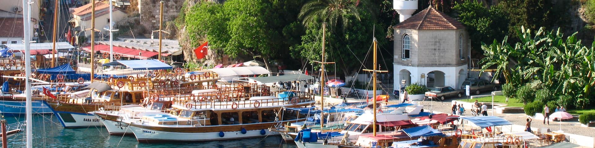 A marina with boats docked, a small white building with a tower, and a stone wall in the background.