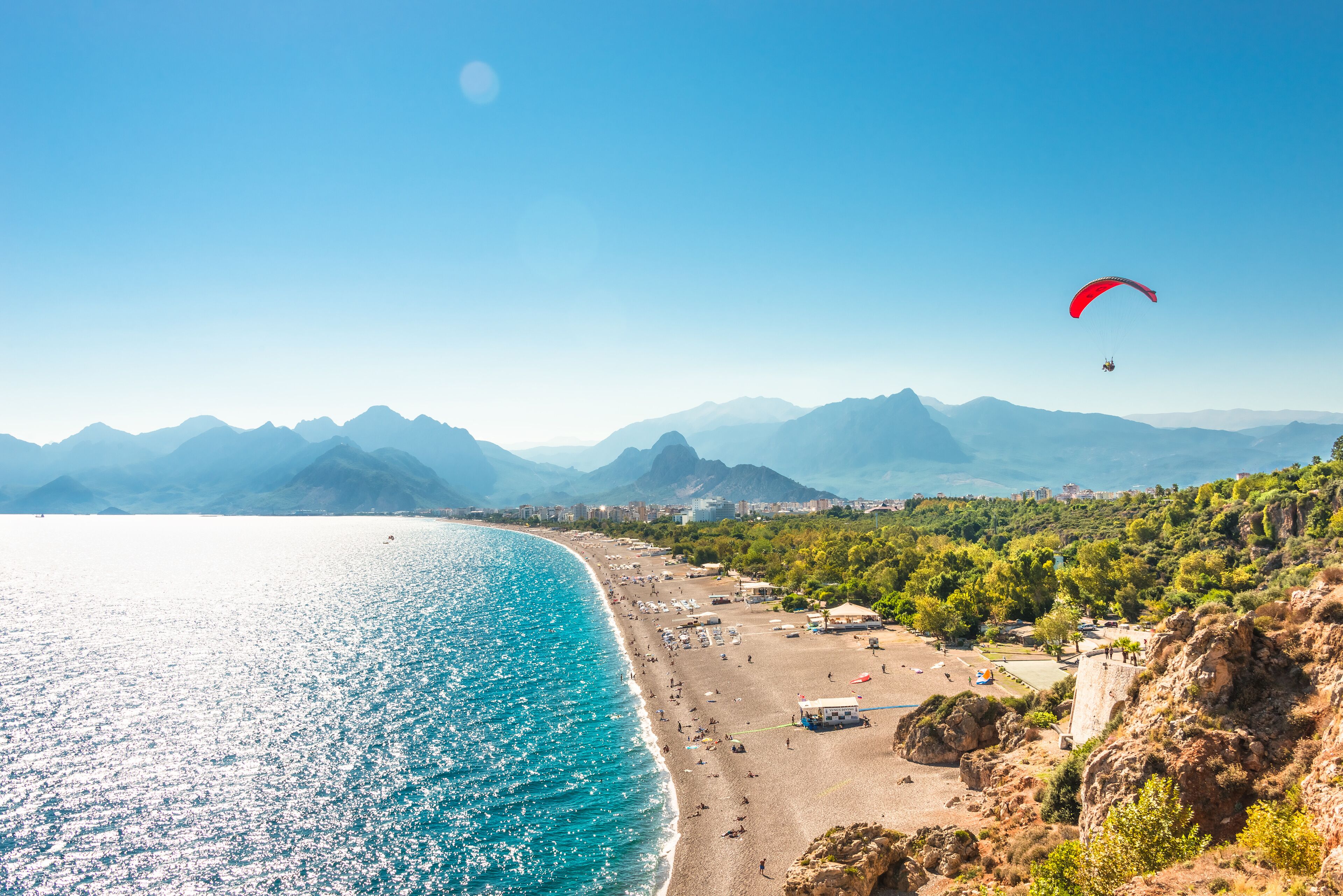 A coastal beach with a paraglider, clear blue sea, and mountains in the background.