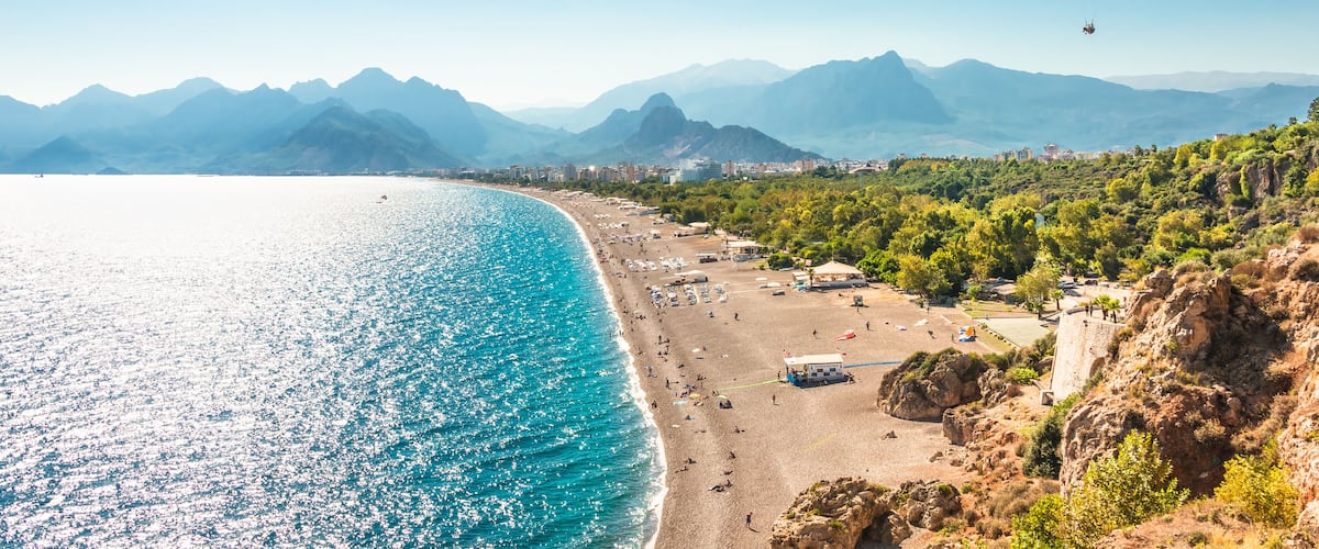 A coastal beach with a paraglider, clear blue sea, and mountains in the background.