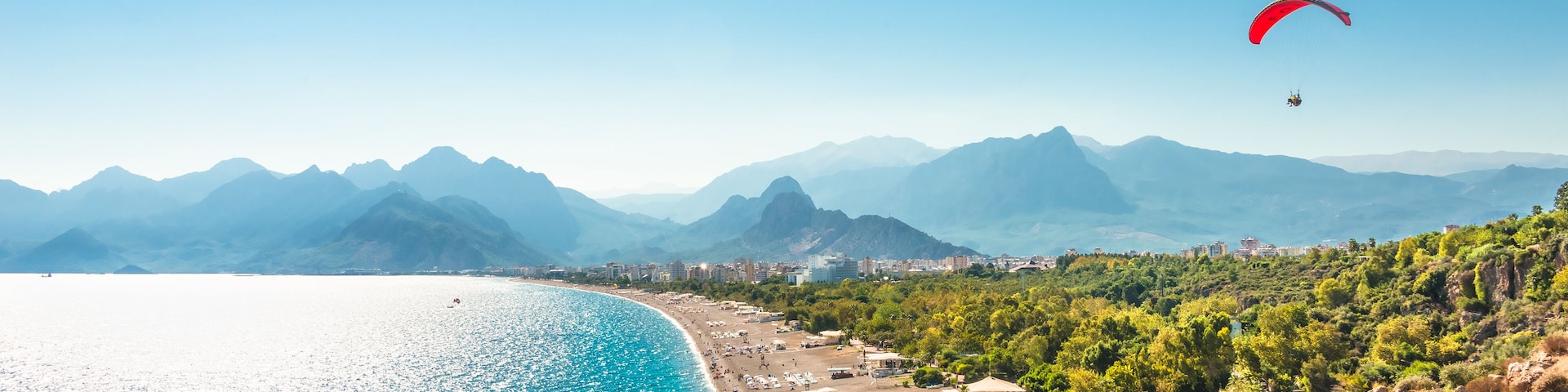 A coastal beach with a paraglider, clear blue sea, and mountains in the background.