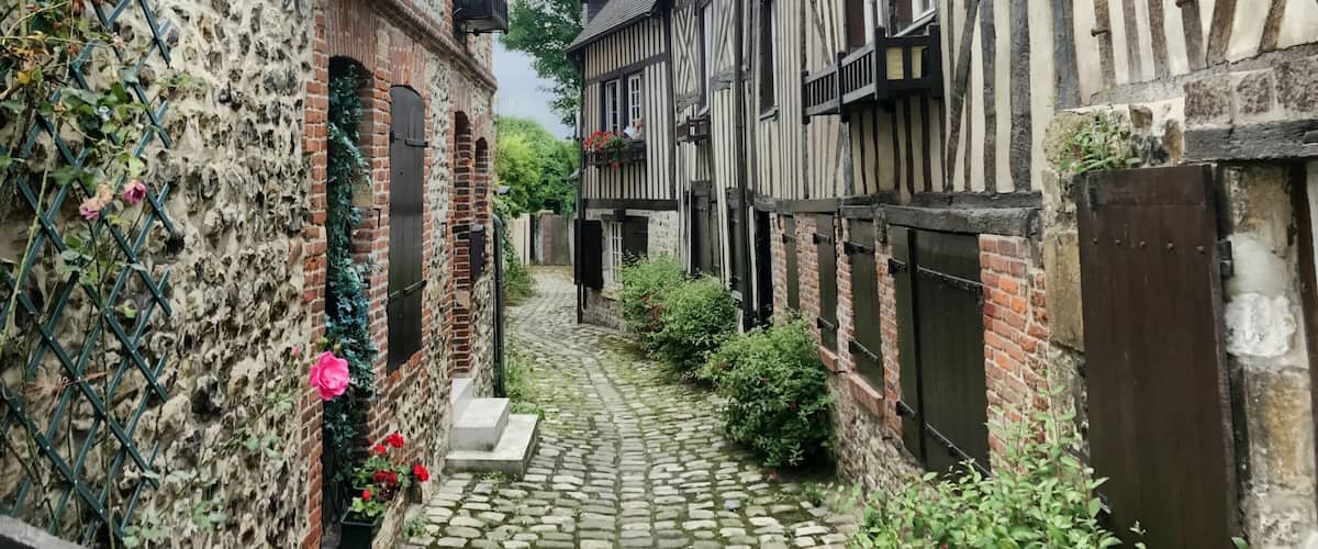 A narrow cobblestone alley flanked by half-timbered buildings with brick walls and flowering plants.