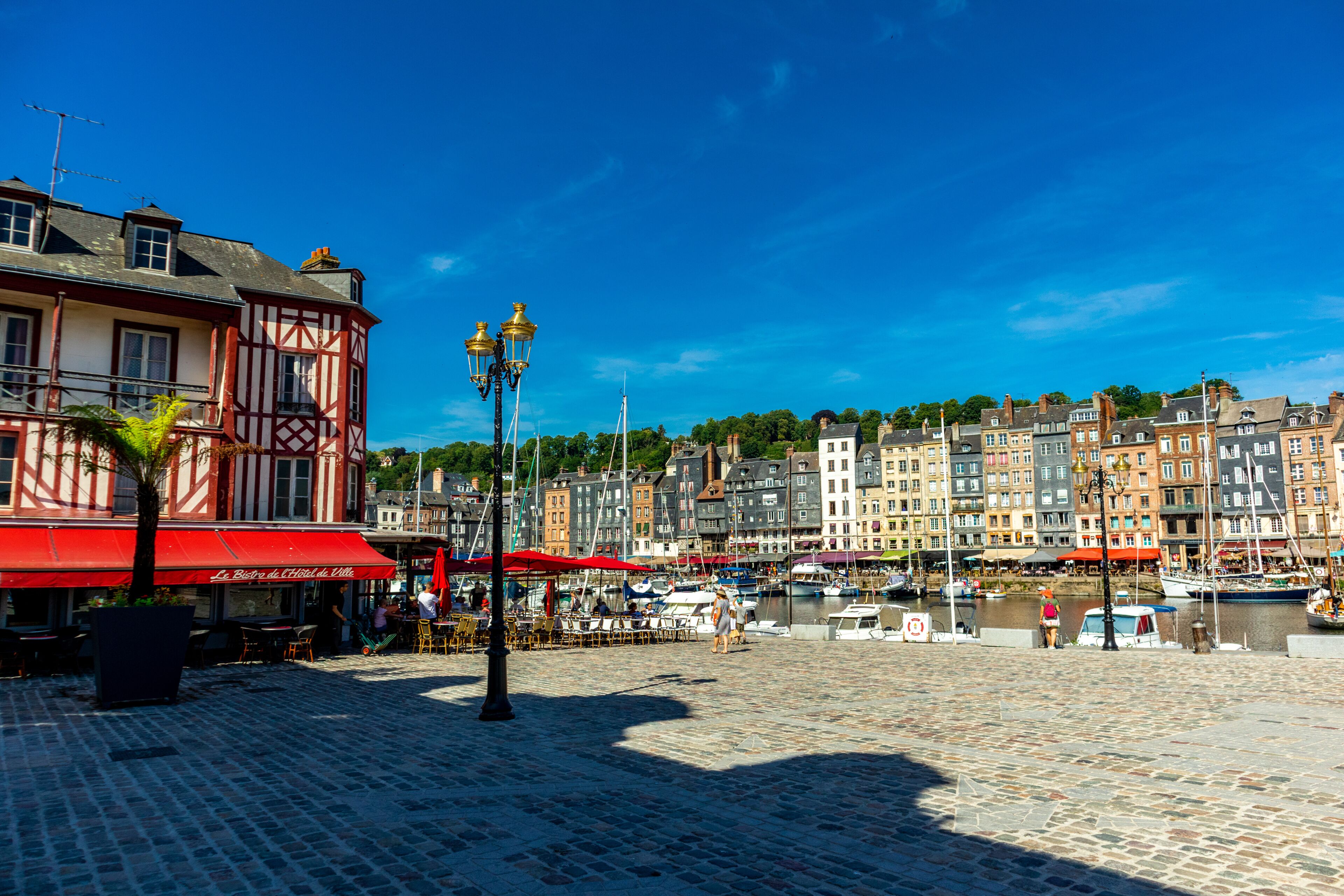 A cobblestone waterfront with historic buildings, a café with red awnings, and boats docked in the background.