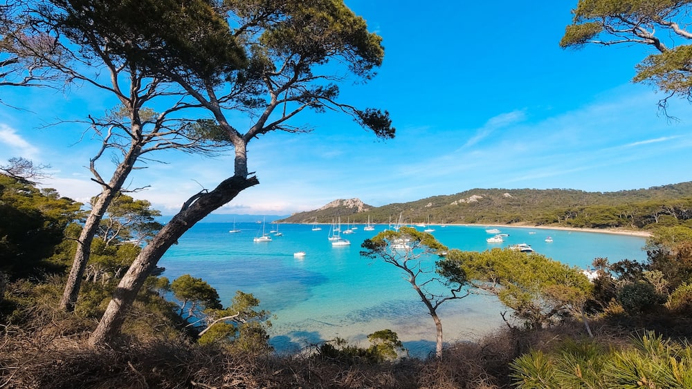 Un paysage côtier avec une eau turquoise cristalline, des bateaux amarrés et, en arrière-plan, des collines.