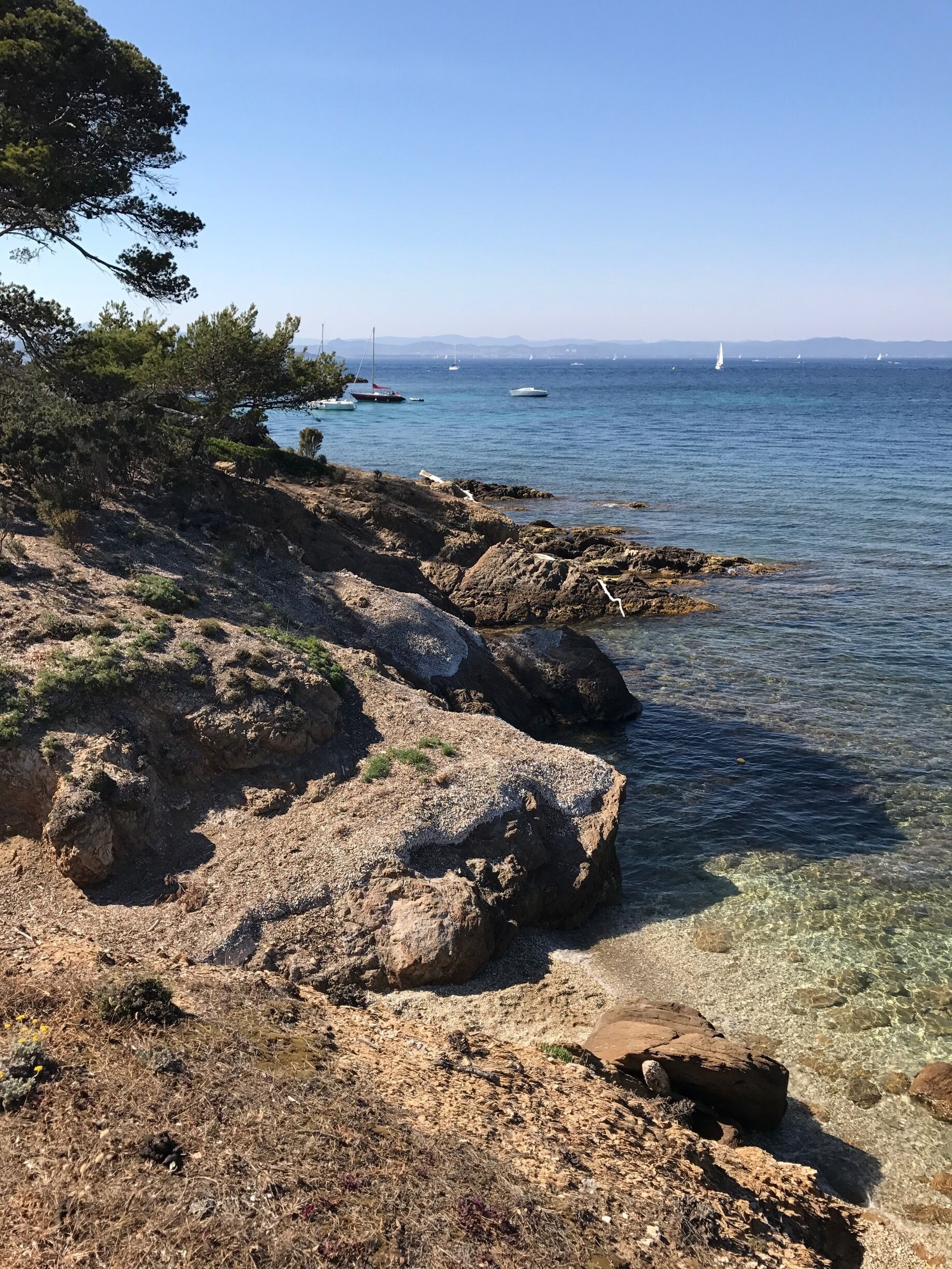 Una costa rocosa con agua cristalina, barcos a lo lejos y un cielo despejado.