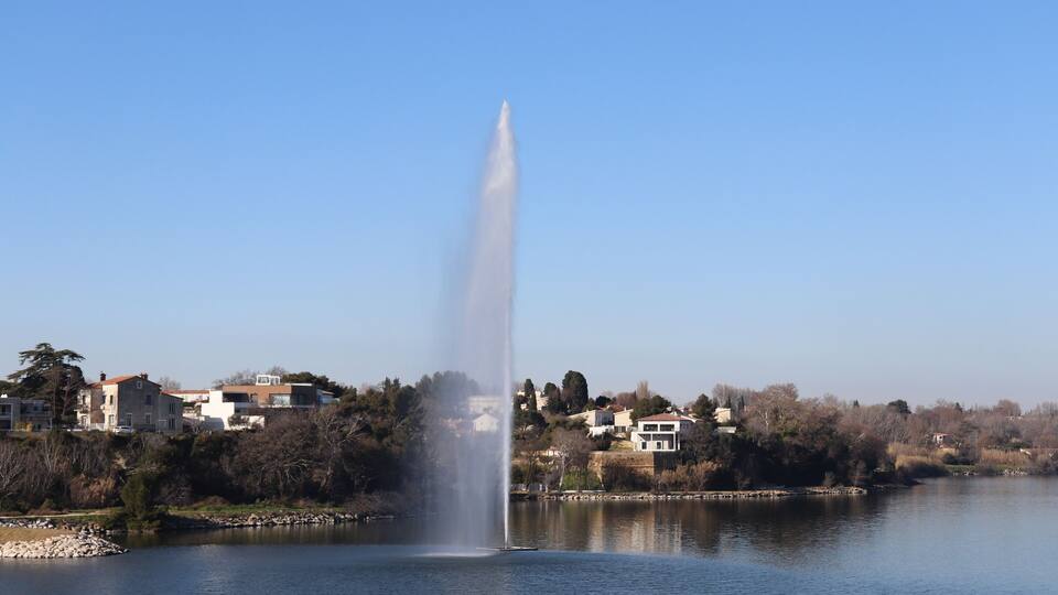 A fountain jetting water into the air, surrounded by a calm lake, with houses and trees in the background.