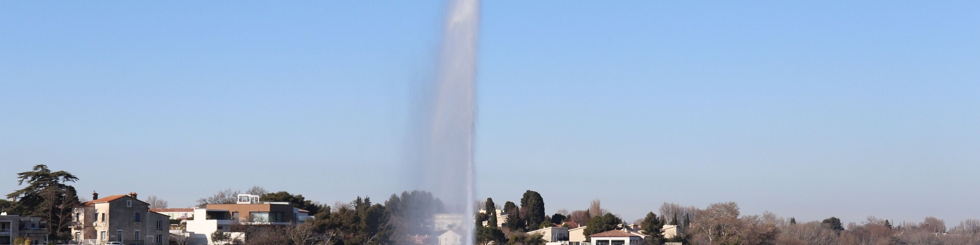 A fountain jetting water into the air, surrounded by a calm lake, with houses and trees in the background.