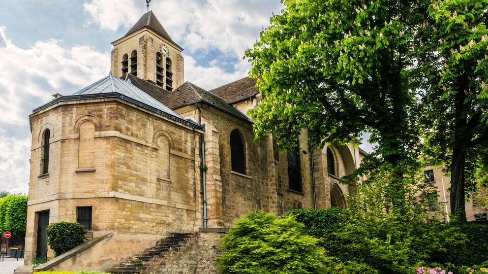 A historic stone church with a bell tower, surrounded by lush greenery and flowering plants.