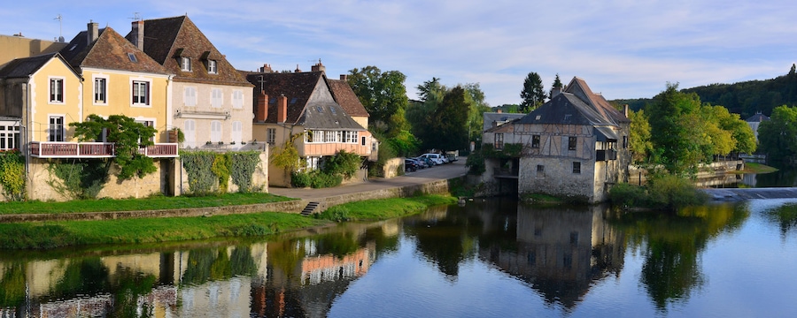 Une rivière paisible, bordée de maisons dont les façades se reflètent dans l’eau, le tout entouré de verdure et d’arbres.