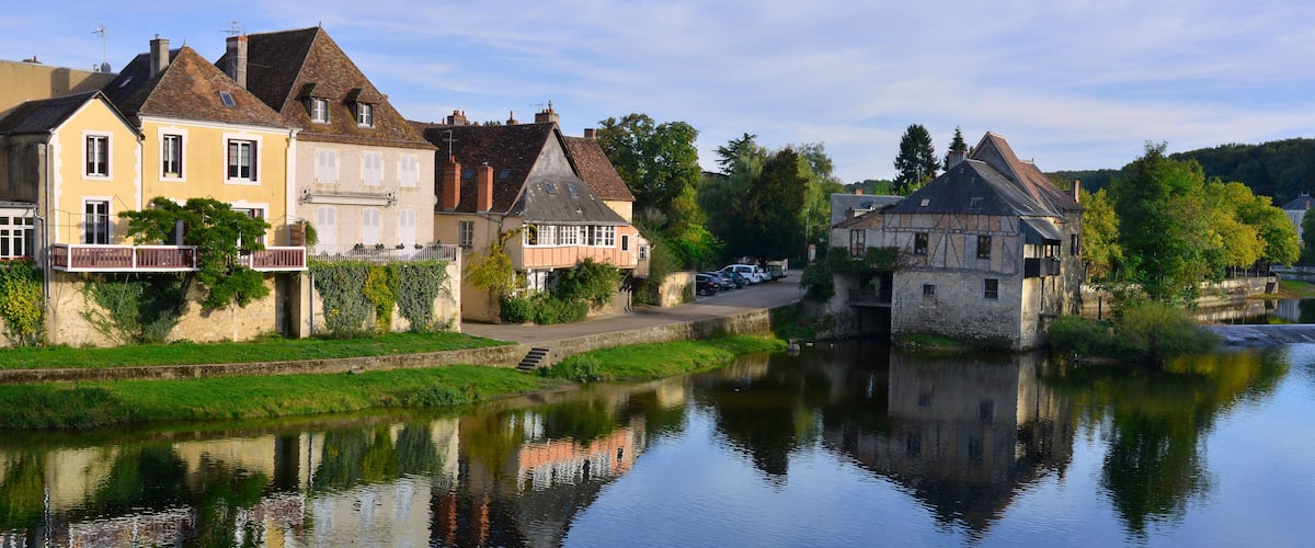 A calm river with houses reflecting on the water, surrounded by greenery and trees.