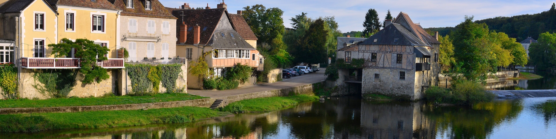 A calm river with houses reflecting on the water, surrounded by greenery and trees.