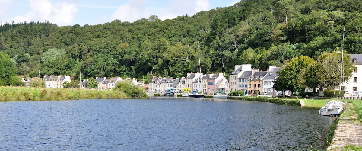 A river with houses along the banks, a boat docked on the right, and a forested hill in the background.